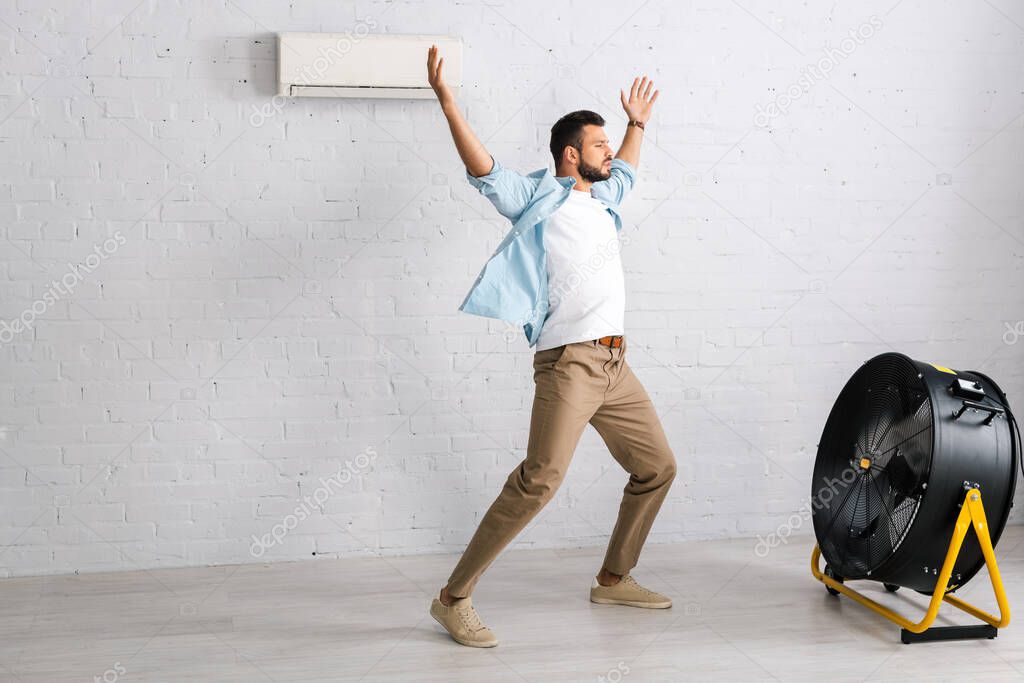 Handsome man standing with raised hands near electric fan and air conditioner at home 