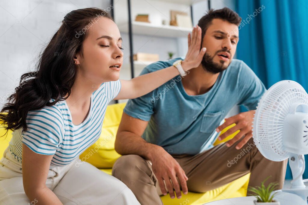 Selective focus of young woman moving away boyfriend while sitting near electric fan