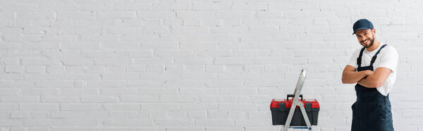 Panoramic shot of smiling workman looking at camera near toolbox on ladder and white wall