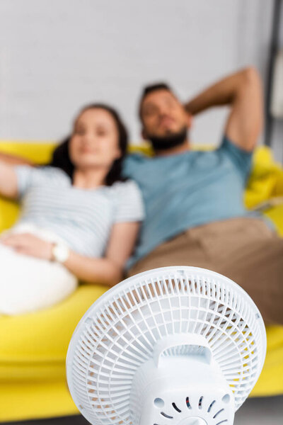 Selective focus of electric fan near couple sitting on sofa at home 