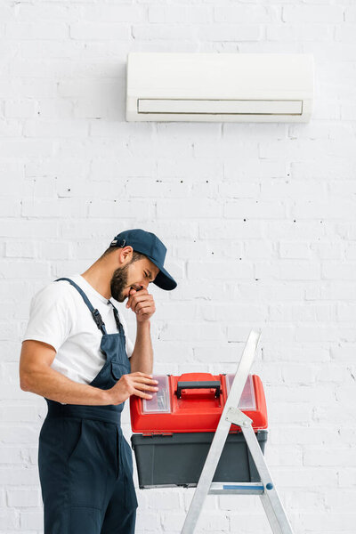 Injured workman with finger near mouth opening toolbox on ladder near air conditioner on wall 