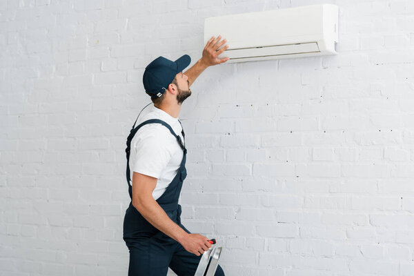Side view of workman in uniform holding screwdriver while fixing air conditioner on wall
