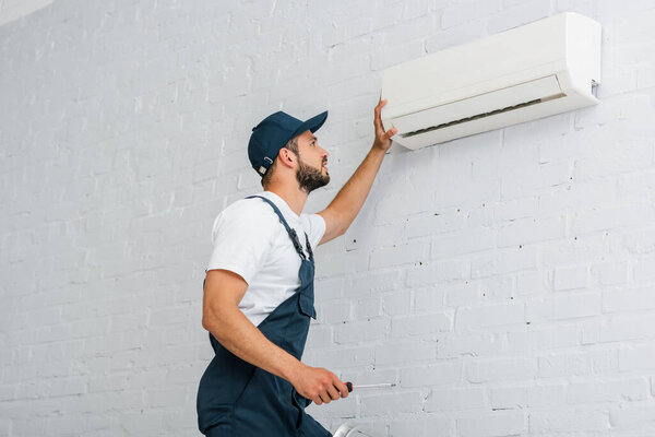 Side view of workman in workwear holding screwdriver near air conditioner 