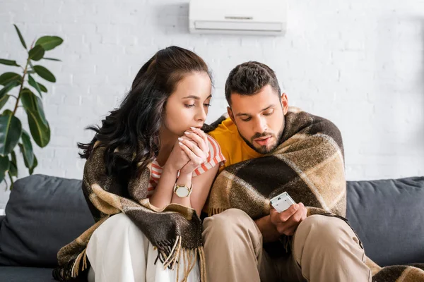 裹着格子花的女人坐在男朋友身边 把冷气机遥控器放在沙发上 — 图库照片