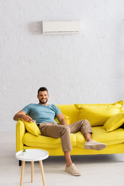 Handsome man smiling at camera while holding remote controller of air conditioner at home — Stock Image Handsome Man Smiling Camera While Holding Remote Controller Air Conditioner Royalty Free Stock Photos