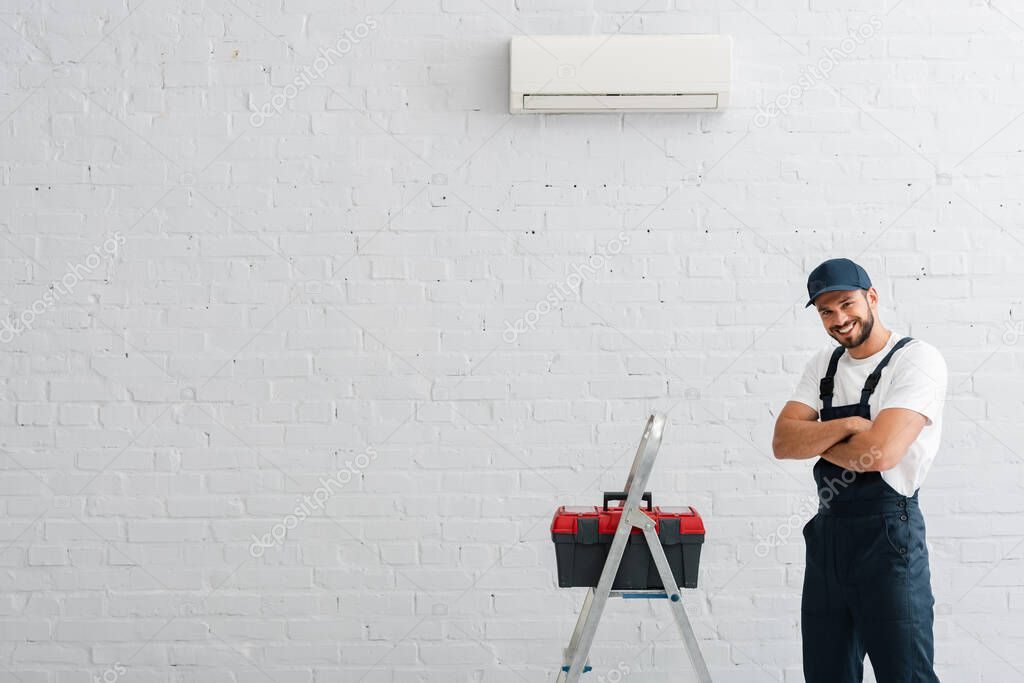 Smiling workman with crossed arms looking at camera near toolbox on ladder and air conditioner on wall
