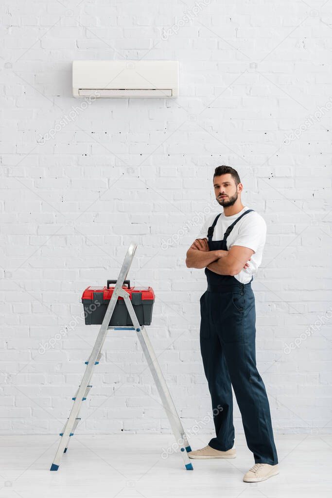 Handsome handyman looking at camera near toolbox on ladder and air conditioner on white wall