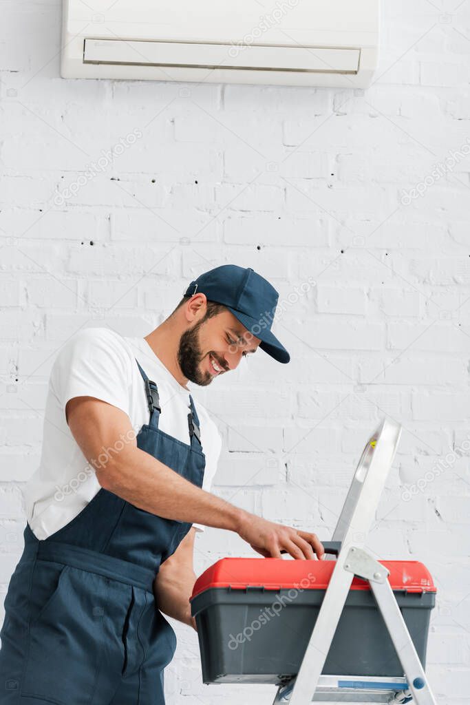 Smiling workman in uniform holding toolbox near ladder and air conditioner on white wall