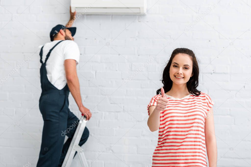 Selective focus of smiling woman showing thumb up while handyman fixing air conditioner 