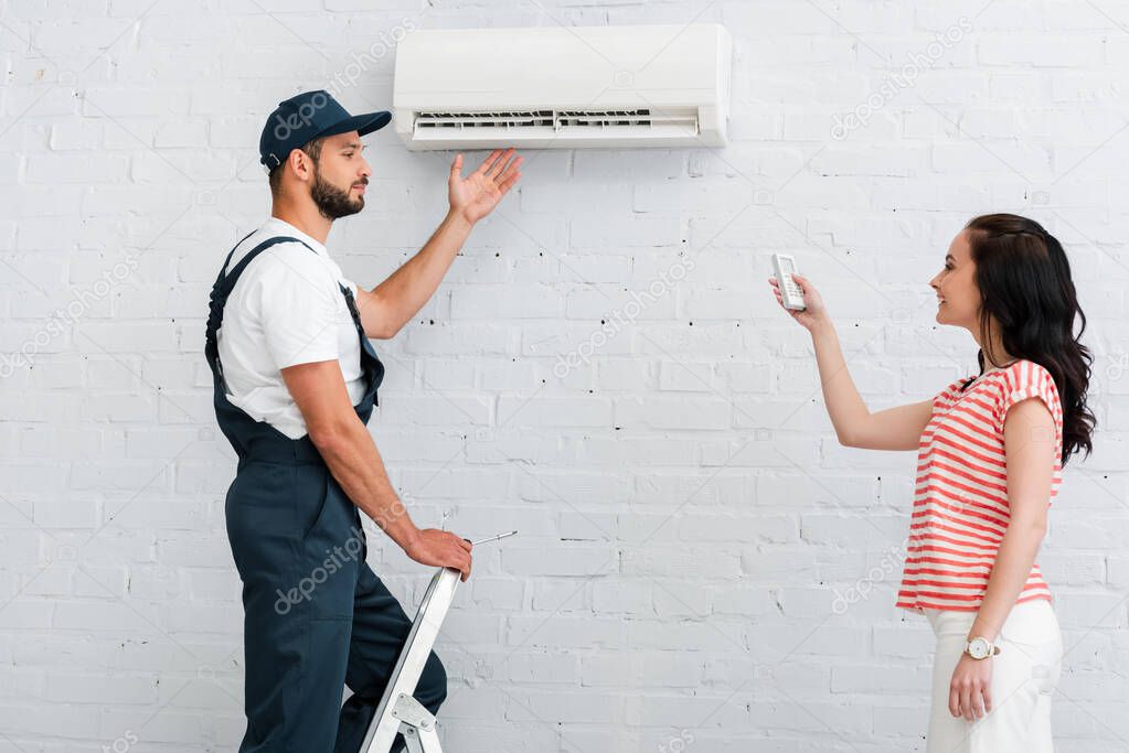 Side view of workman pointing at air conditioner near smiling woman holding remote controller