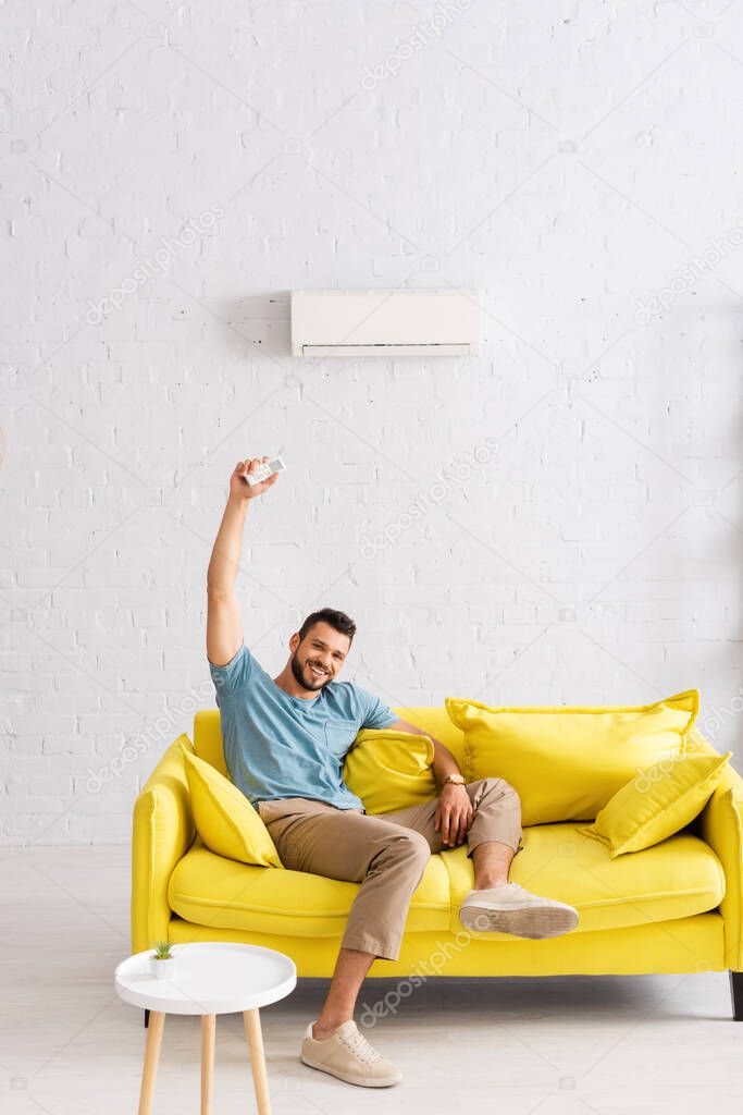 Cheerful man smiling at camera while holding remote controller of air conditioner in living room