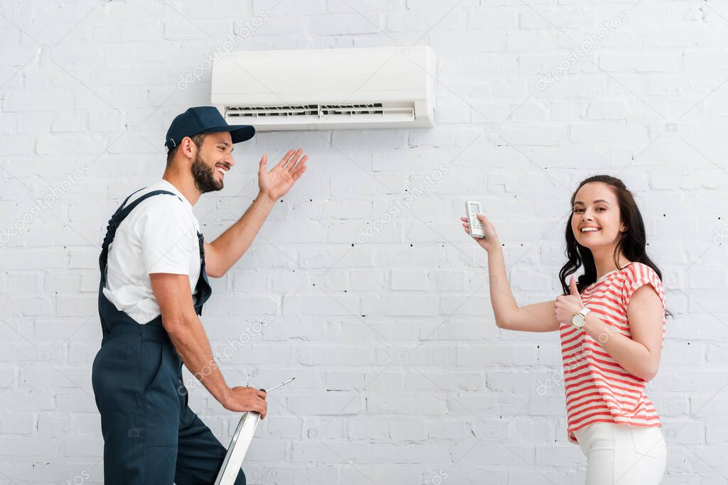 Side view of smiling woman with remote controller of air conditioner showing thumb up near workman in overalls on ladder 
