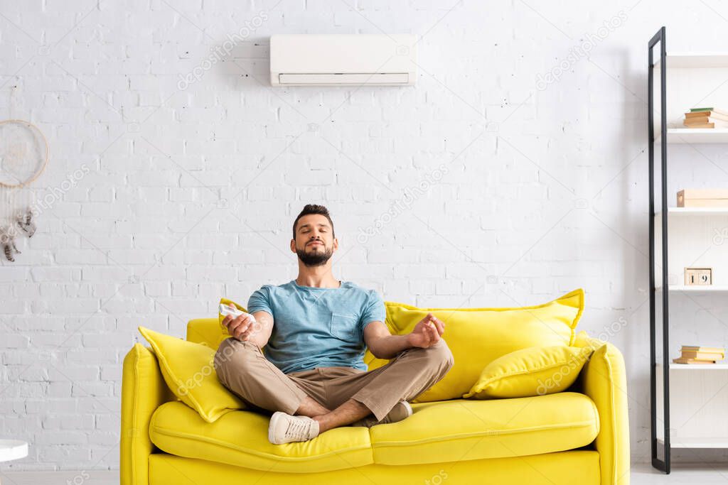 Young man holding remote controller of air conditioner while meditating on sofa 
