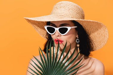 Güneş gözlüklü şık bir kadın ve palmiye yaprağının yanındaki hasır şapkalı turuncu tenli. 