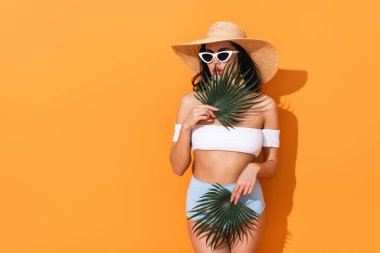 Mayolu şık bir kadın, güneş gözlüğü ve elinde palmiye yaprakları tutan hasır bir şapka.
