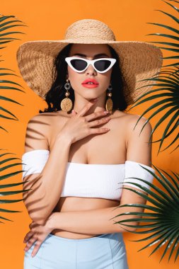 Mayolu, güneş gözlüklü ve hasır şapkalı çekici bir kız. Palmiye yapraklarının yanında boyna dokunuyor.