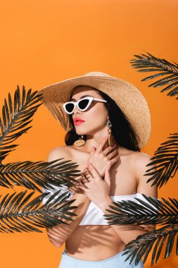 Güneş gözlüklü, hasır şapkalı, palmiye yapraklarının yanında boyna dokunan şık bir kadın.
