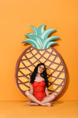 attractive and barefoot woman in swimsuit and sunglasses sitting in lotus pose near paper cut pineapple on orange, summer concept  clipart