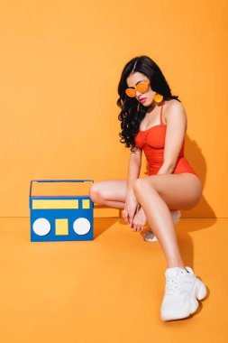 Spor ayakkabılı, mayolu ve güneş gözlüklü şık bir kadın. Turuncu üzerine kağıt kesiği olan müzik kutusunun yanında oturuyor.
