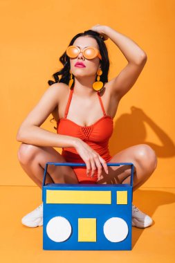 Spor ayakkabılı şık bir kız, mayo ve güneş gözlüğü kağıdın yanında oturuyor müzik kutusunu kesiyor ve turuncu saçlarına dokunuyor.