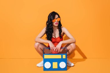 Spor ayakkabılı, mayo giyen ve güneş gözlüklü modaya uygun genç bir kadın. Kağıt kesikli, turuncu bir müzik kutusunun yanında oturuyor.