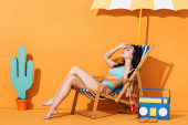 atraktivní žena v plavkách dotýkající se slunečních brýlí a sedící na lehátku u koktejlu, žabky, papírová krabice a kaktus na oranžové