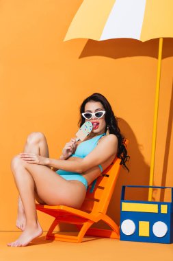 Güneş gözlüklü ve mayo giyen modaya uygun bir kadın portakalda dondurma yalarken kağıt teyp ve şemsiyenin yanında oturuyor.