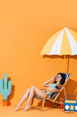 Güneş gözlüklü ve mayo giyen modaya uygun bir kadın kokteyl, terlik, kağıt teyp, kaktüs ve şemsiye yanında oturuyor.