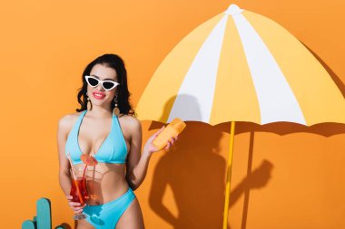 Mayolu ve güneş gözlüklü mutlu kadın elinde güneş kremi ve turuncu üzerine kağıt şemsiye yanında kokteyl tutuyor.
