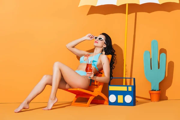 スタイリッシュなサングラスと水着を身につけた女の子がオレンジのカクテルを飲みながら机の上に座っています — ストック写真