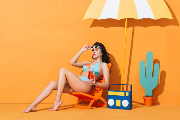 戴着太阳镜 身穿泳衣的时髦女人坐在纸箱和伞旁边的甲板椅子上 拿着橙子鸡尾酒 — 图库照片