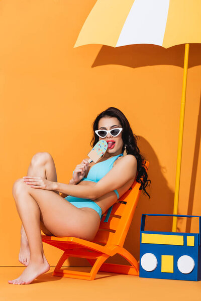 trendy woman in sunglasses and swimwear sitting on deck chair near paper boombox and umbrella while licking ice cream on orange