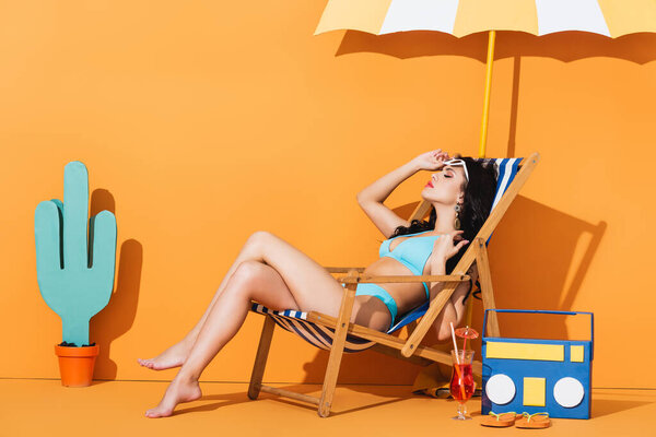 attractive woman in swimwear touching sunglasses and sitting on deck chair near cocktail, flip flops, paper boombox and cactus on orange