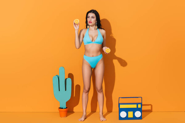 woman in bathing suit holding orange halves while standing near paper cactus and boombox on orange 