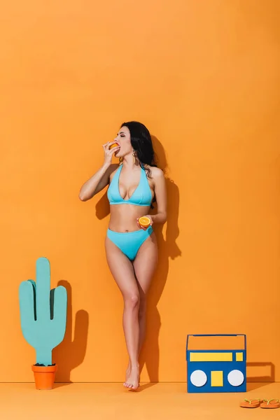 Frau Badeanzug Isst Orange Während Sie Der Nähe Von Papierkaktus — Stockfoto