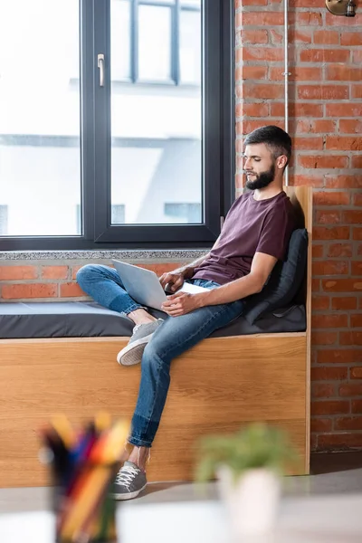 selective focus of bearded businessman sitting on window bench and using laptop