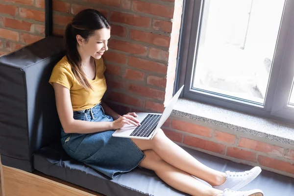 happy businesswoman sitting on window bench and using laptop in office