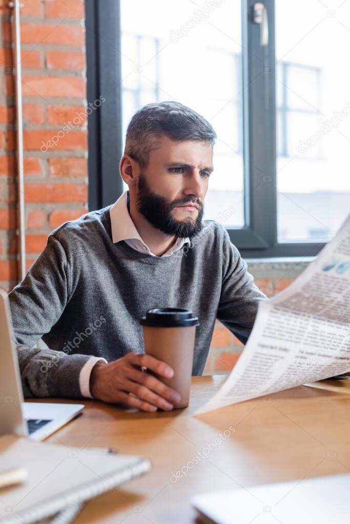 selective focus of bearded businessman holding coffee to go and reading newspaper