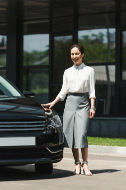 Şehir sokağında arabanın yanında kameraya gülümseyen çekici bir iş kadını. 