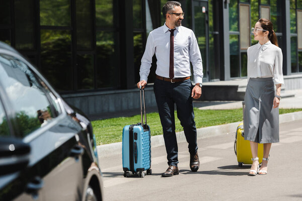 Selective focus of businessman with suitcase smiling at colleague on urban street 