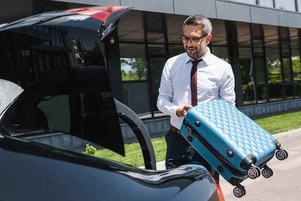 Selective focus of smiling businessman putting suitcase in car truck on urban street