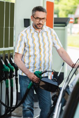 Yakıt ikmal istasyonunda arabanın yanında yakıt ikmali yaparken kameraya bakan yakışıklı adamın seçici odağı.