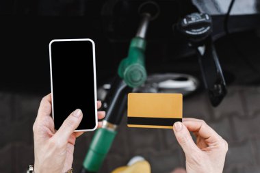 Kadın elinde boş ekran ve kredi kartıyla akıllı telefon tutarken benzin istasyonunda yakıt ikmali yaparken görüntüsü kesilmiş. 