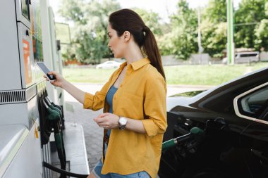 Woman holding smartphone and credit card near auto on car refueling station  clipart