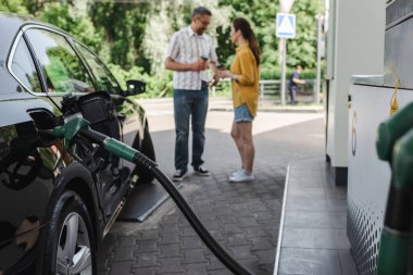 Selective focus of fueling nozzle in gas tank of car and couple holding paper cups on urban street  clipart