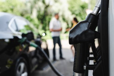 Selective focus of fueling nozzle on gas station and couple standing near auto outdoors  clipart