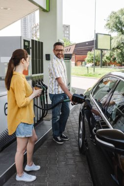 Selective focus of smiling man fueling car near wife with coffee to go on gas station  clipart