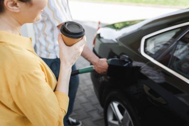 Cropped view of woman holding coffee to go while husband fueling auto on gas station  clipart