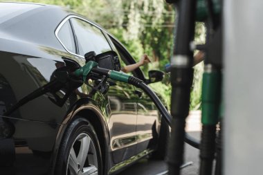 Selective focus of fueling nozzle in gas tank of auto and woman paying with credit card near worker of gas station with payment terminal clipart