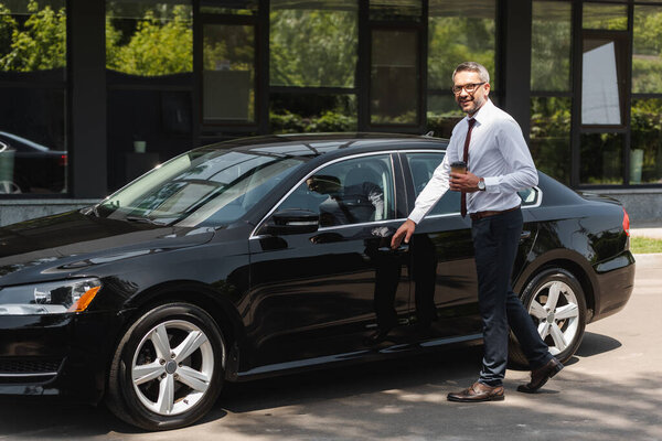 Smiling businessman holding coffee to go near car on urban street 